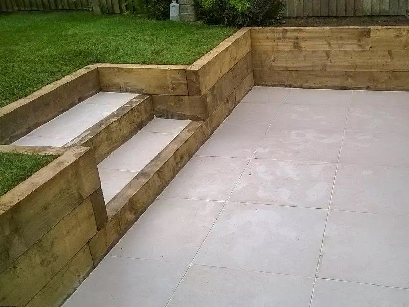 Patio with light-colored square tiles, wood retaining walls, and steps leading up to a grassy area.