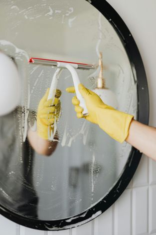 Hands in yellow gloves cleaning a bathroom mirror with a red-and-white squeegee.