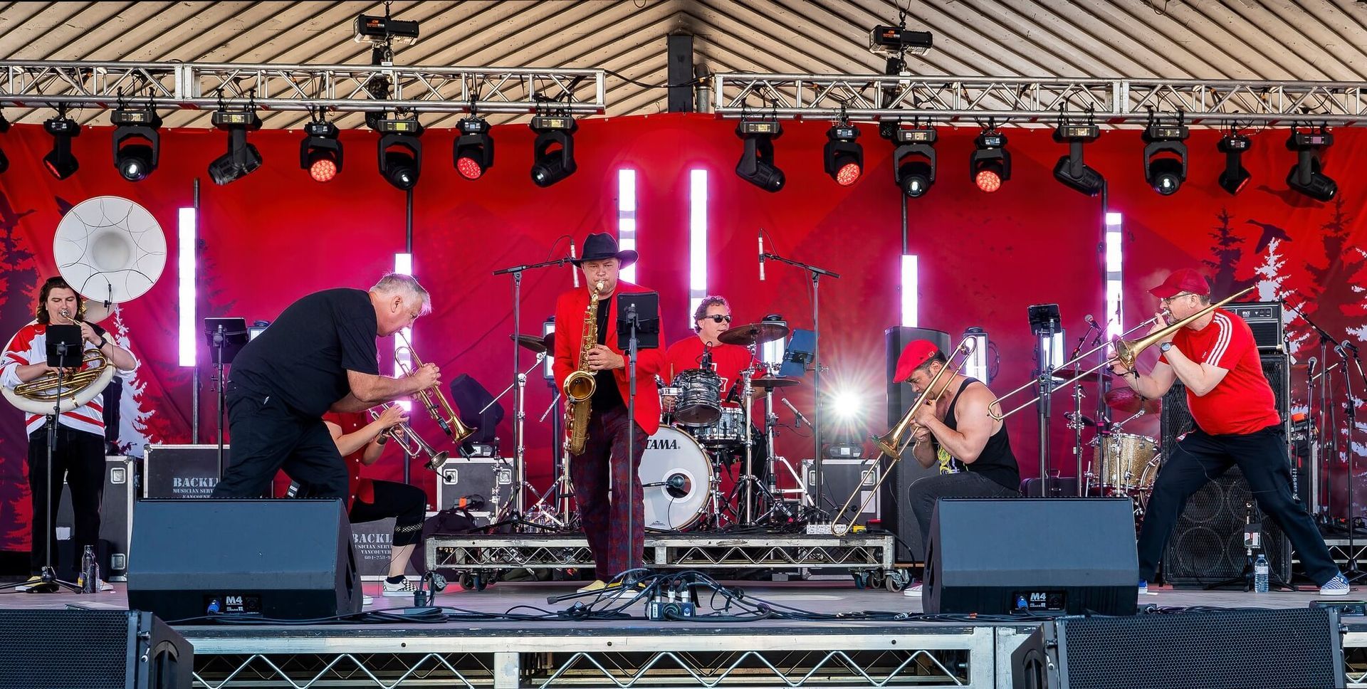 Band performing on an outdoor stage with red backdrop and bright lights