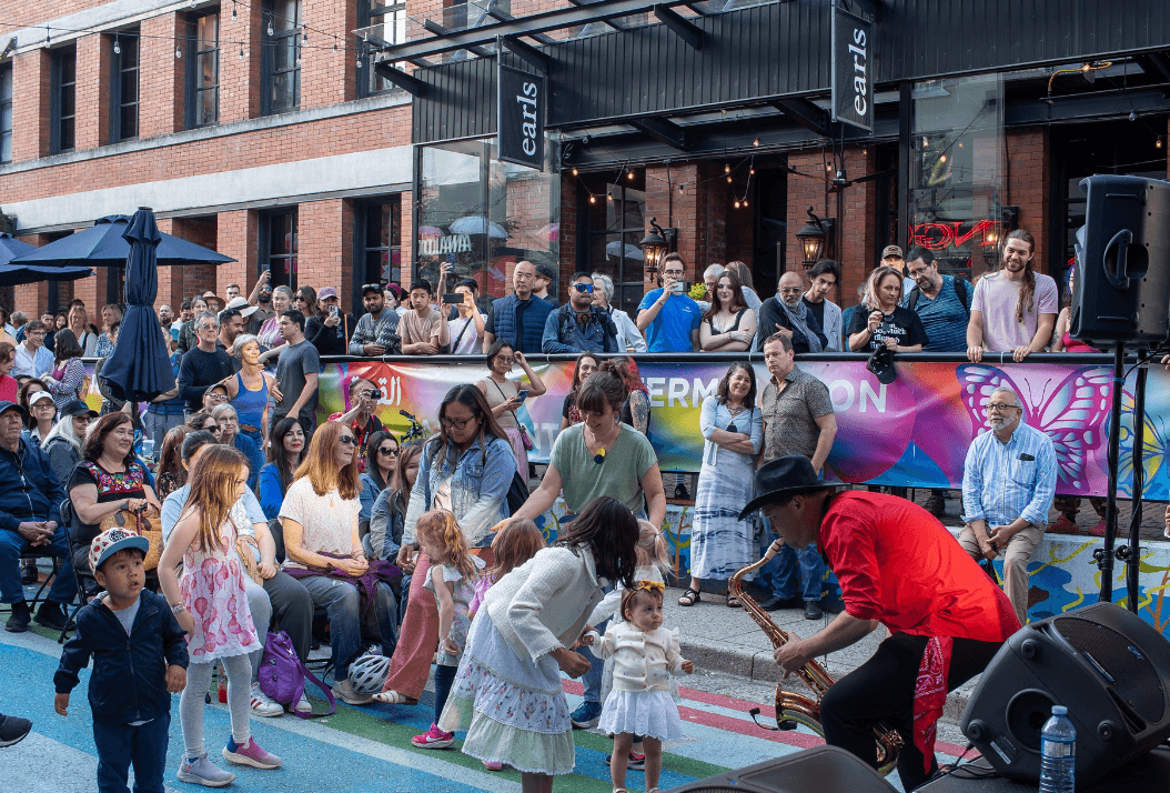 Crowd gathered for an outdoor street performance on a city block, with people seated and standing near a stage.