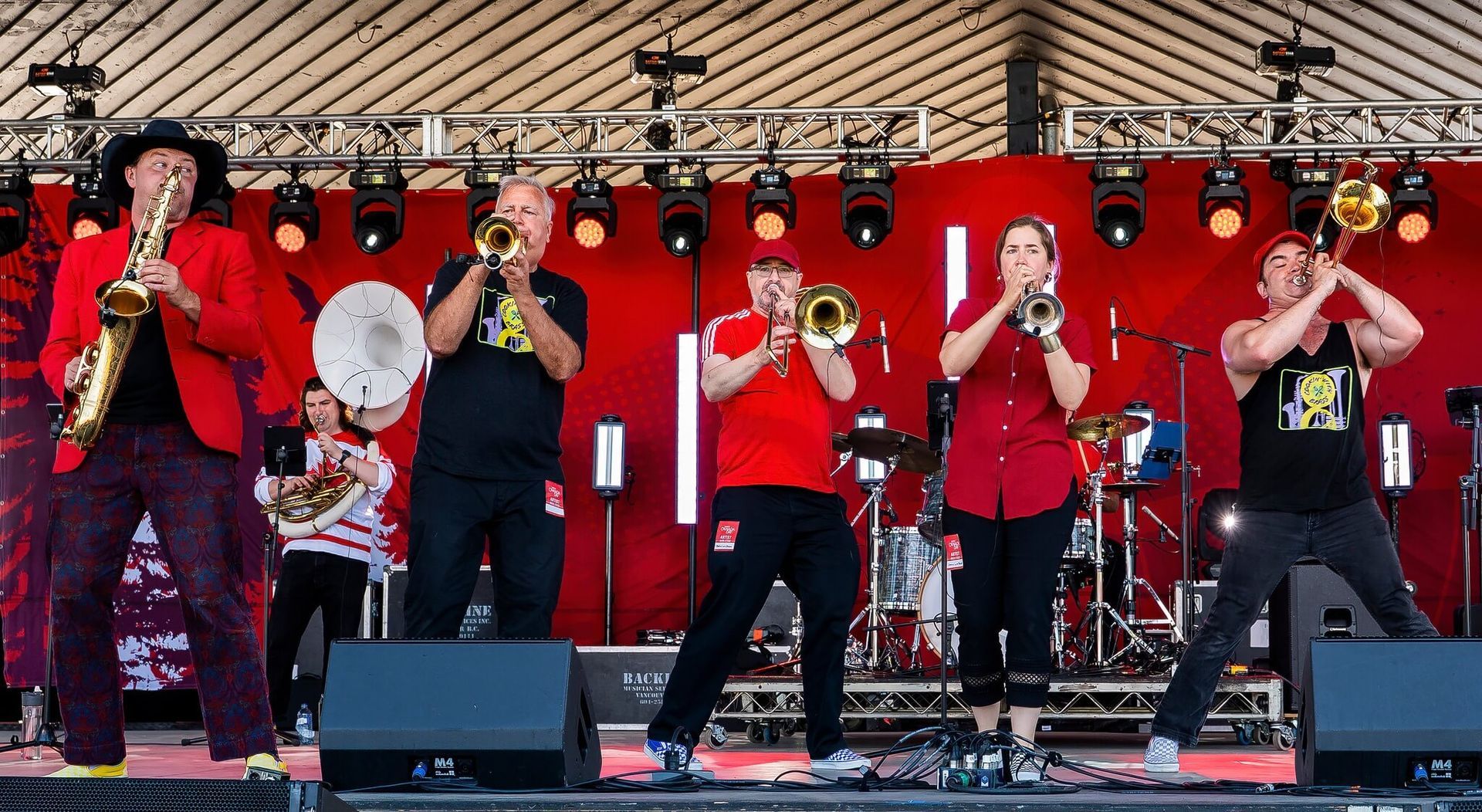 Five-piece brass band in red shirts performing on an outdoor stage with microphones and drums