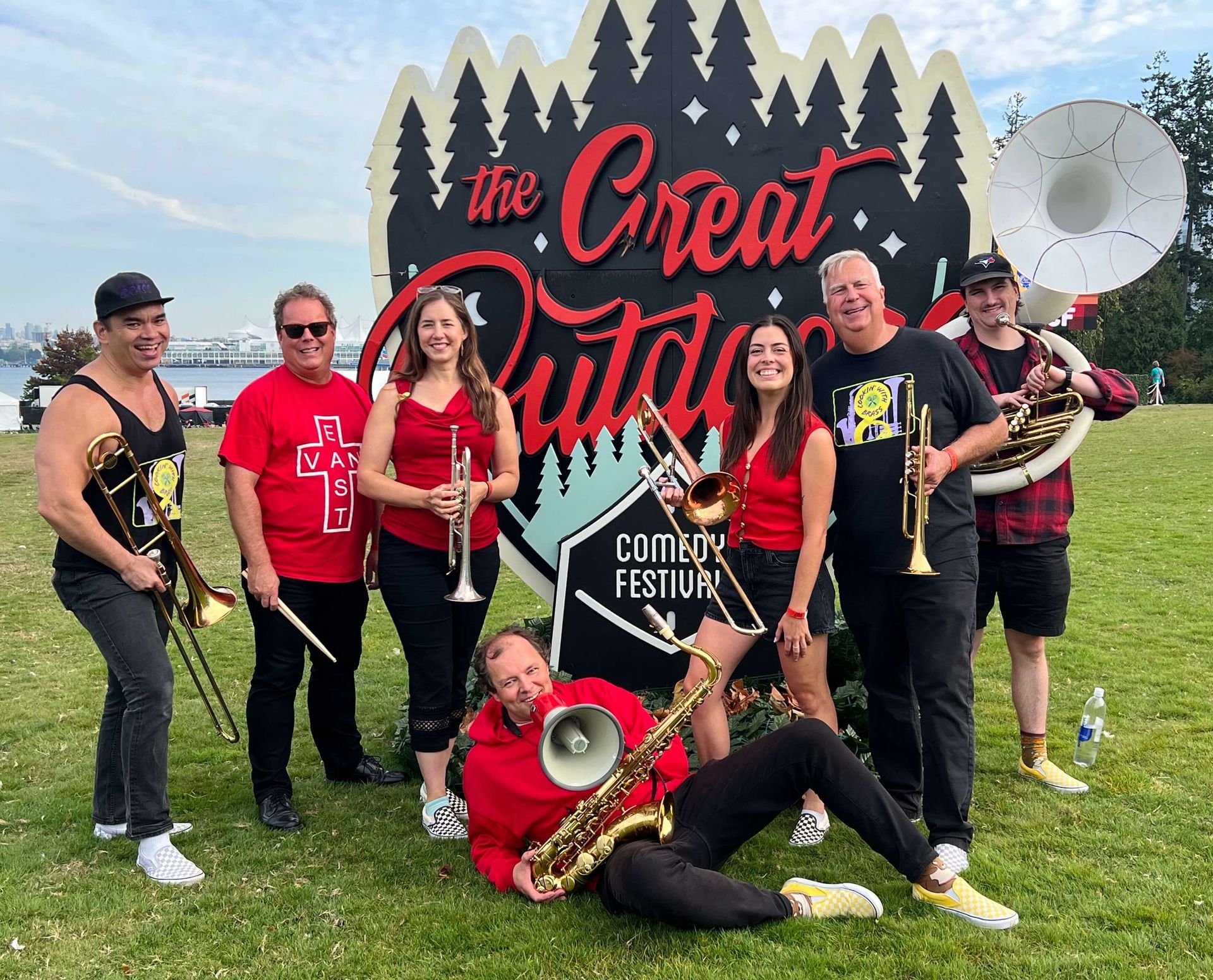 Group posing in front of a “The Great Festa” sign on a grassy field, smiling and holding instruments.