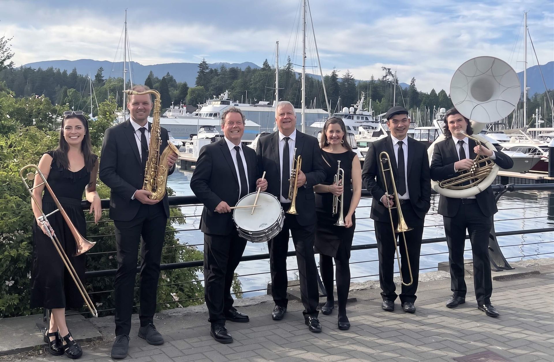 Group of musicians with brass instruments posing on a harbor dock, with boats and water behind them