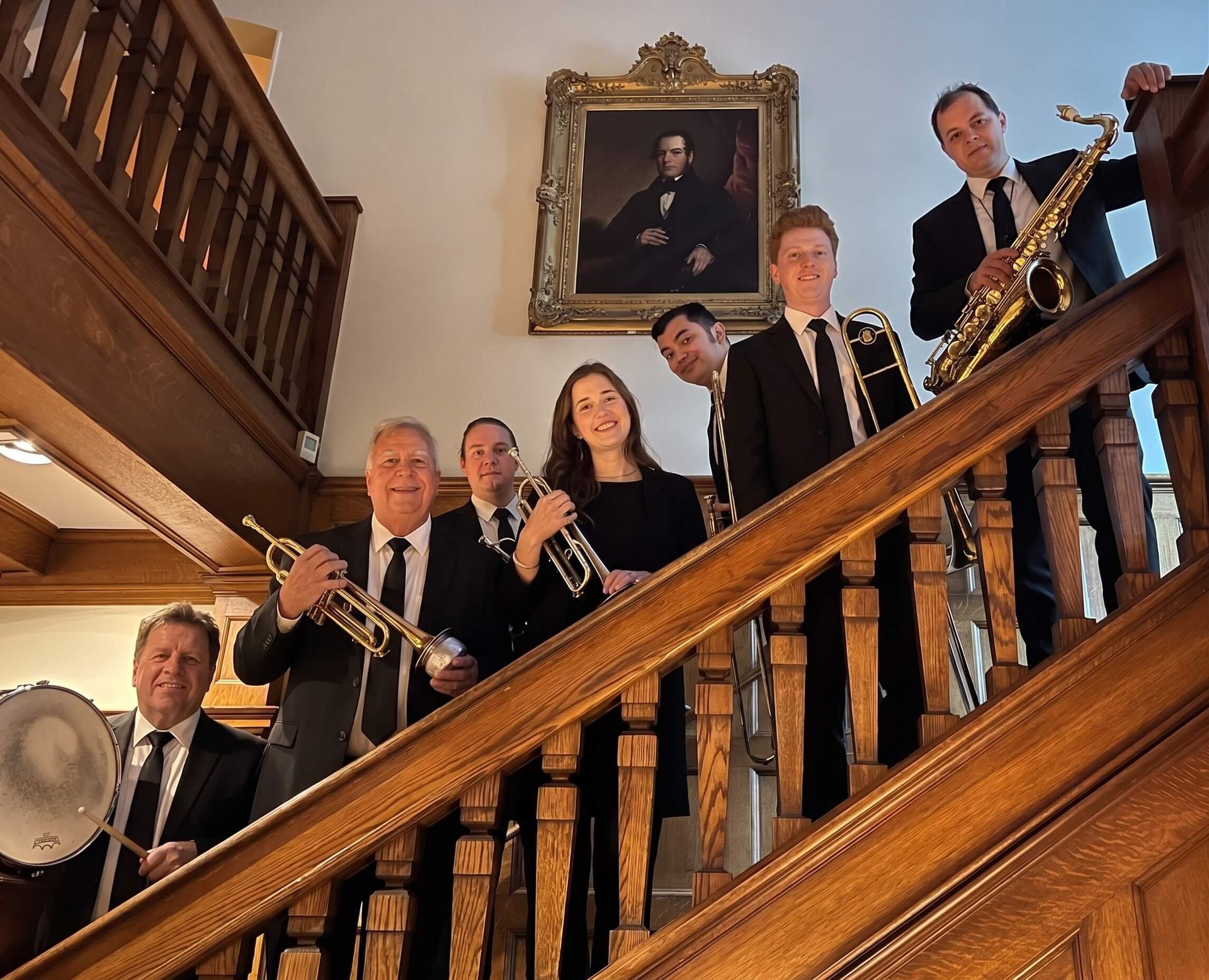 Seven musicians in formalwear pose on a staircase, holding brass instruments in a grand interior.