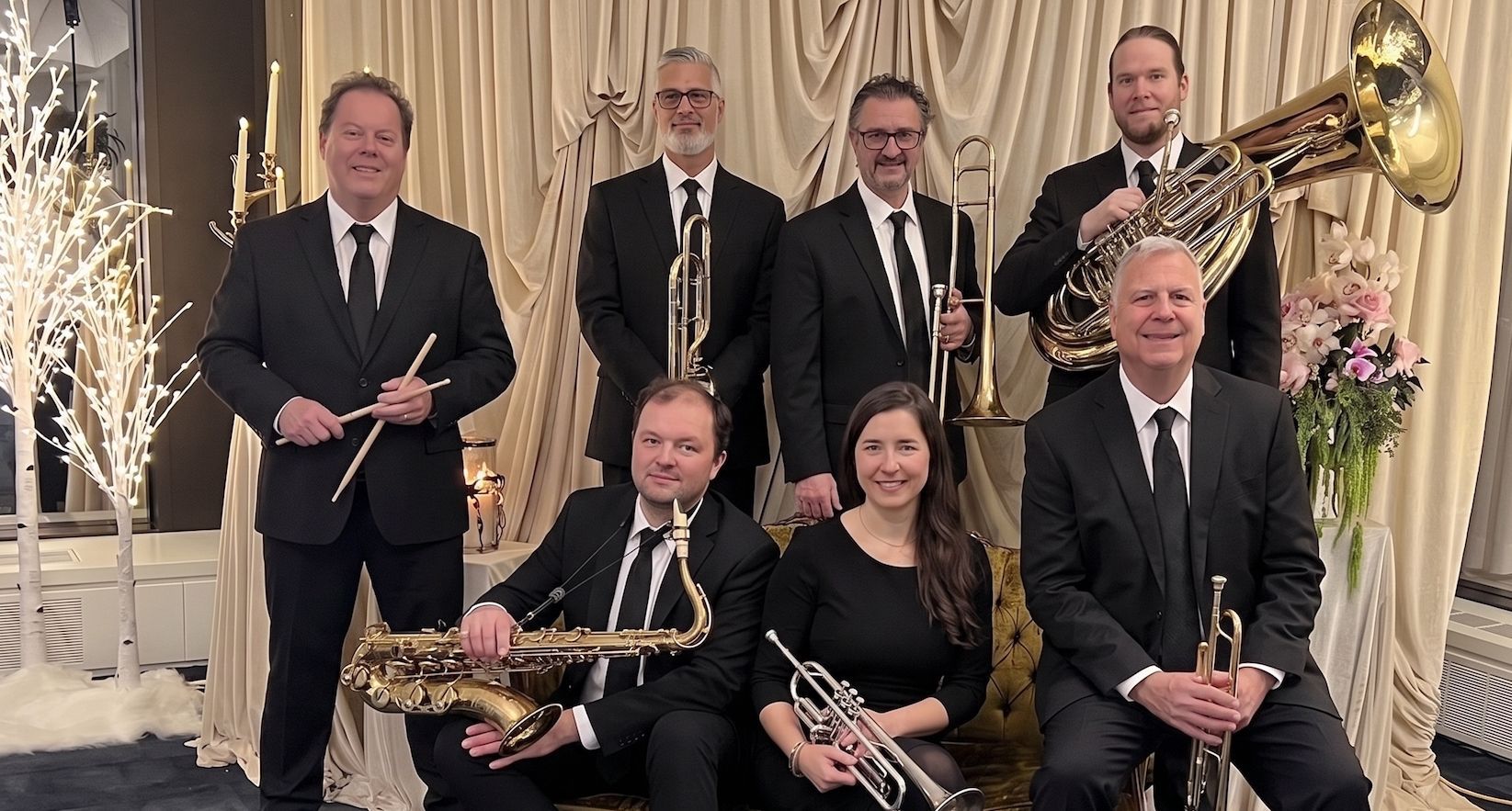 Seven-piece brass ensemble posing indoors in formal black attire, holding brass instruments on a grand staircase.