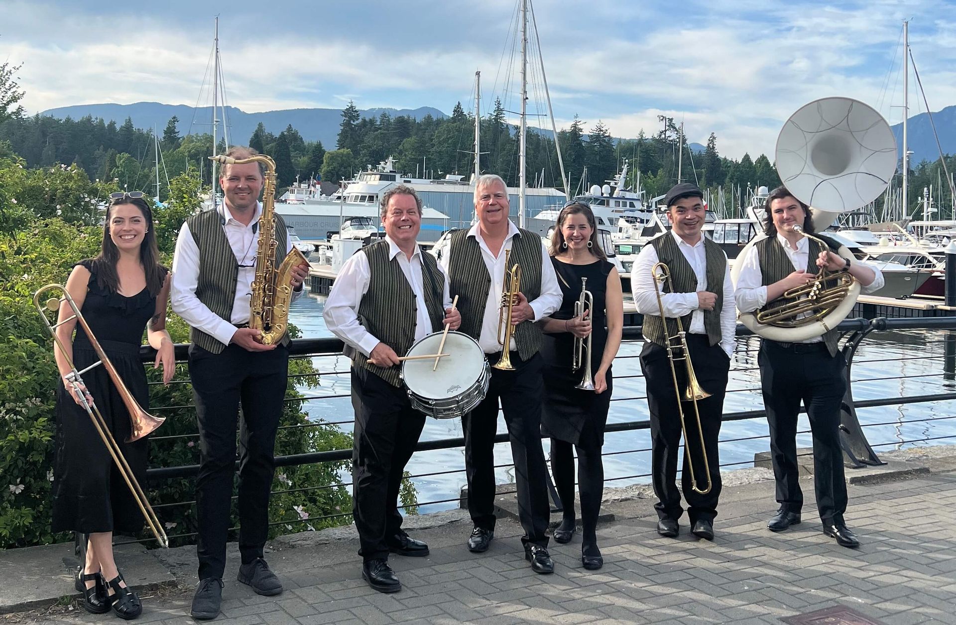 Seven-piece brass band posing outdoors by a harbor, holding instruments with boats and water behind them