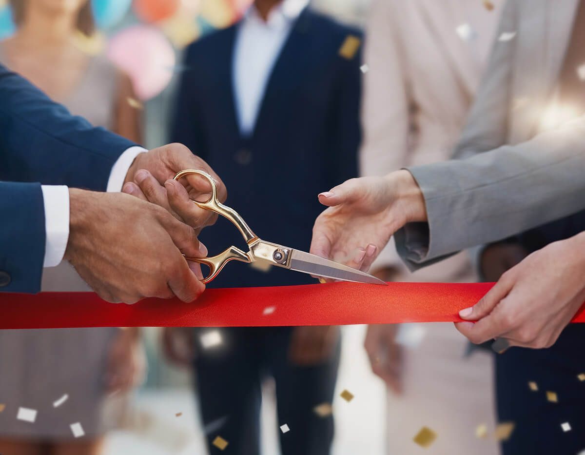 Hands cutting a red ribbon with gold scissors at a ceremony, with confetti falling in the background