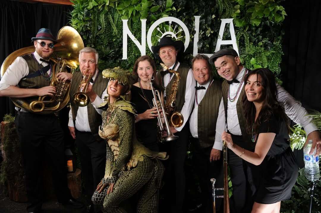 Band posing with saxophones and brass instruments in front of a leafy backdrop with “NOLA” sign