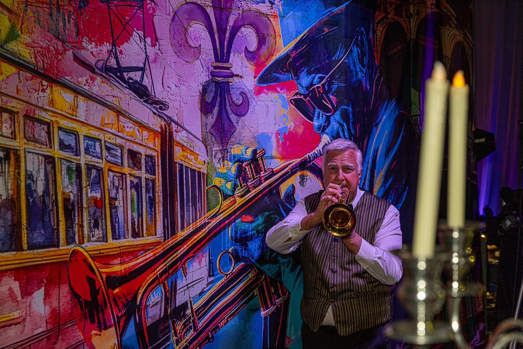 Musician playing trumpet beside a red streetcar, with candles in the foreground and colorful mural backdrop