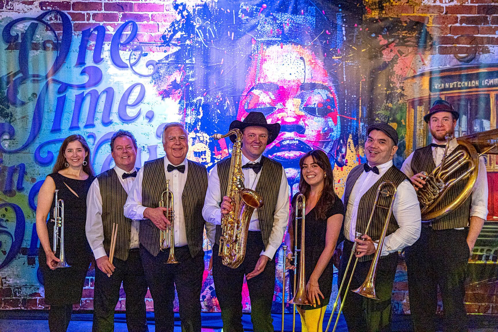 Seven performers in gold costumes stand on stage before a colorful “One Time” backdrop.