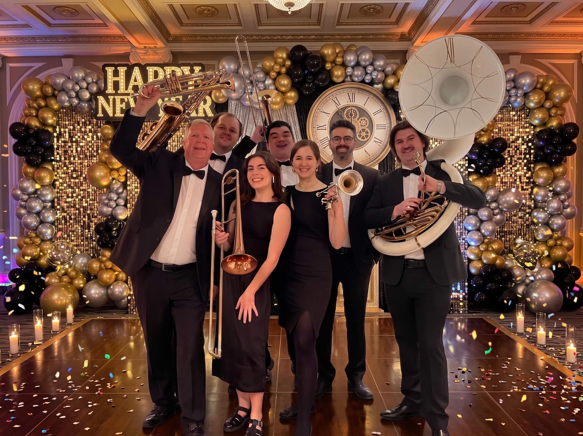 Group of six posing at a glamorous black-and-gold party backdrop with balloons, lights, and confetti