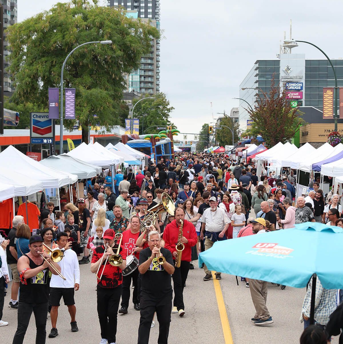 Street parade with marching band and crowds under white tents on a city road