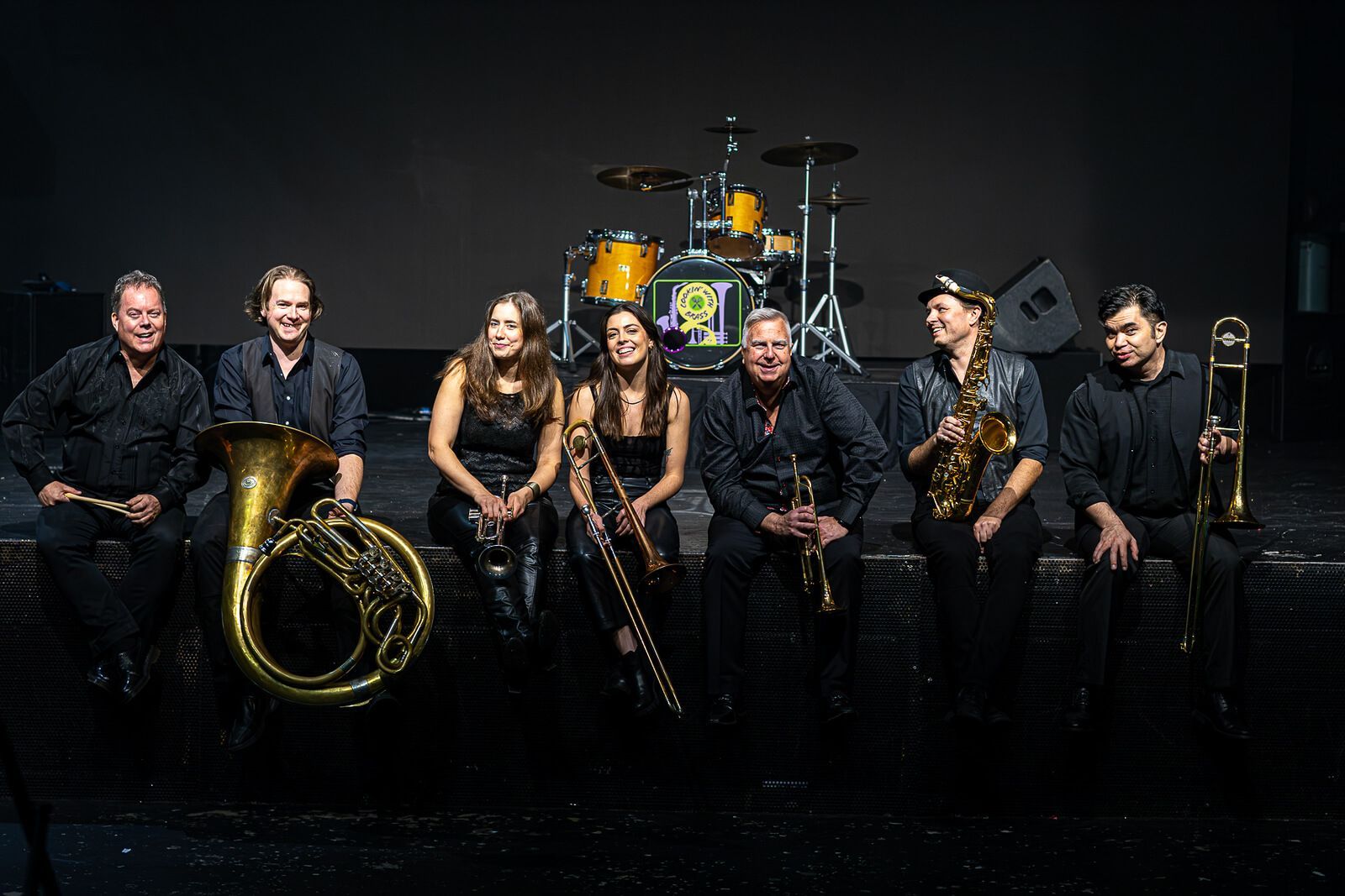 Band group posing in black outfits on a dark stage with drums and brass instruments.