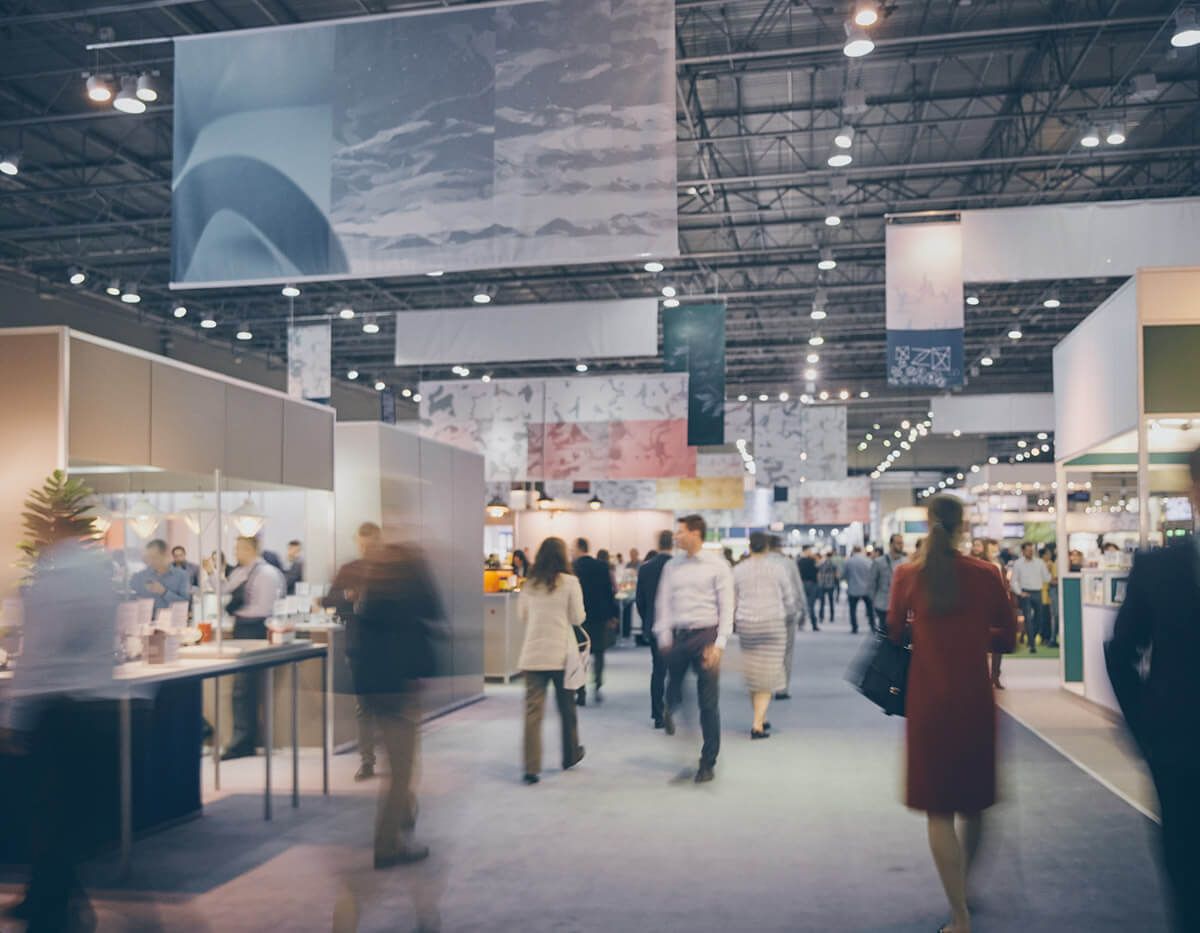 Busy indoor trade show with booths, overhead banners, and people walking through an exhibition hall