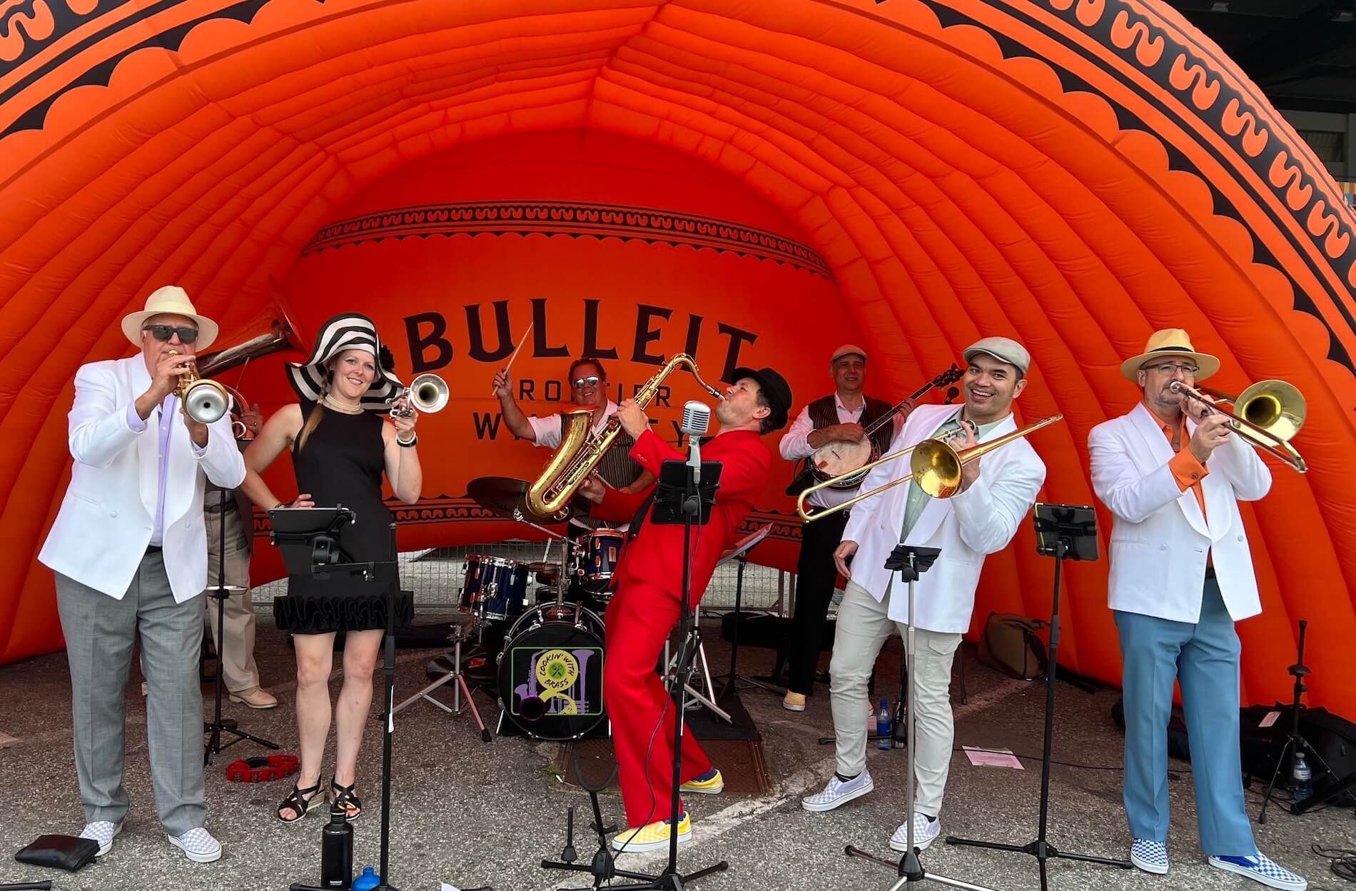 Brass band performing on a red-orange outdoor stage tent, with guitars, saxophone, and trombones.