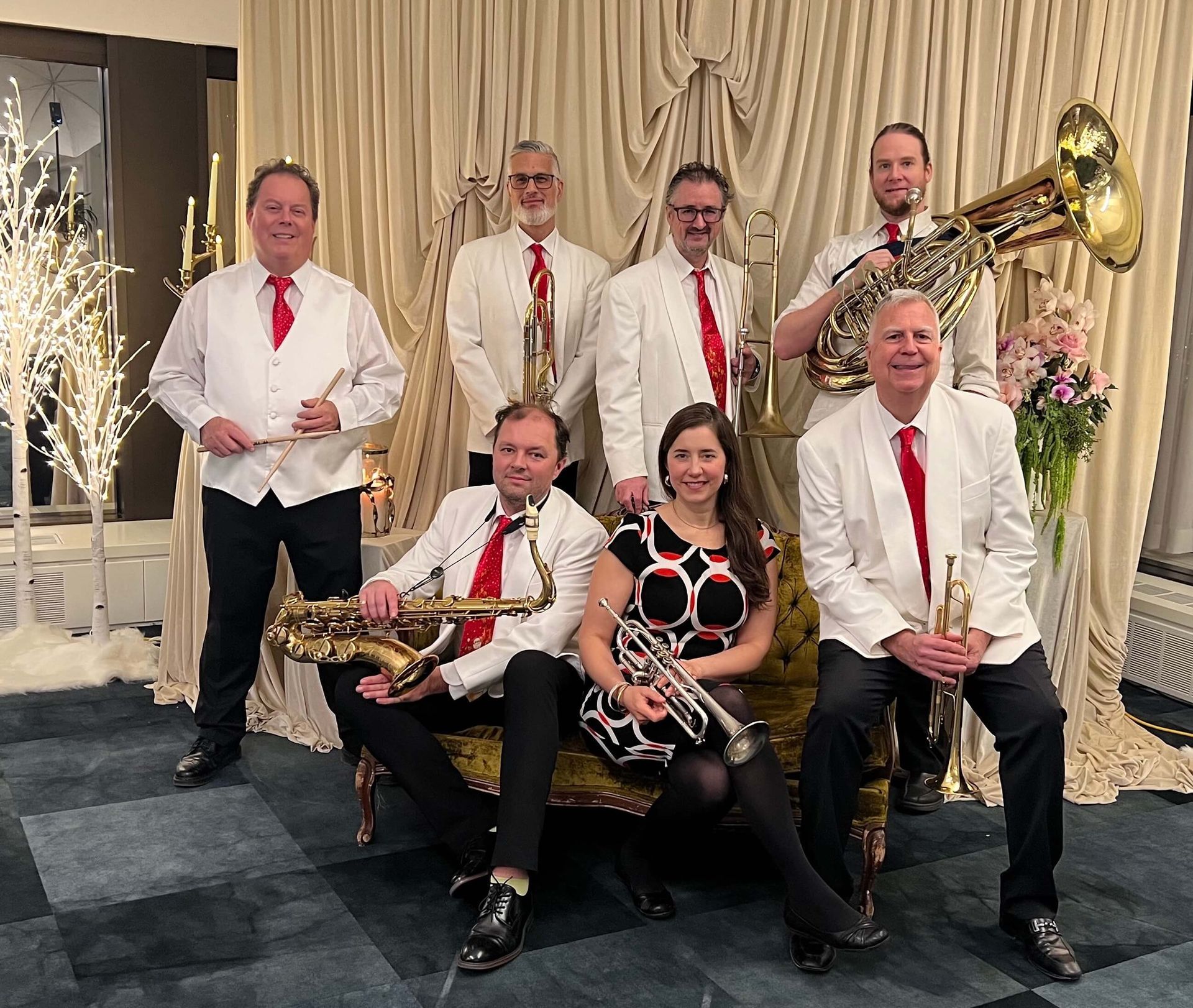 Six-piece brass band in white shirts and red ties posing indoors, with instruments and floral decorations