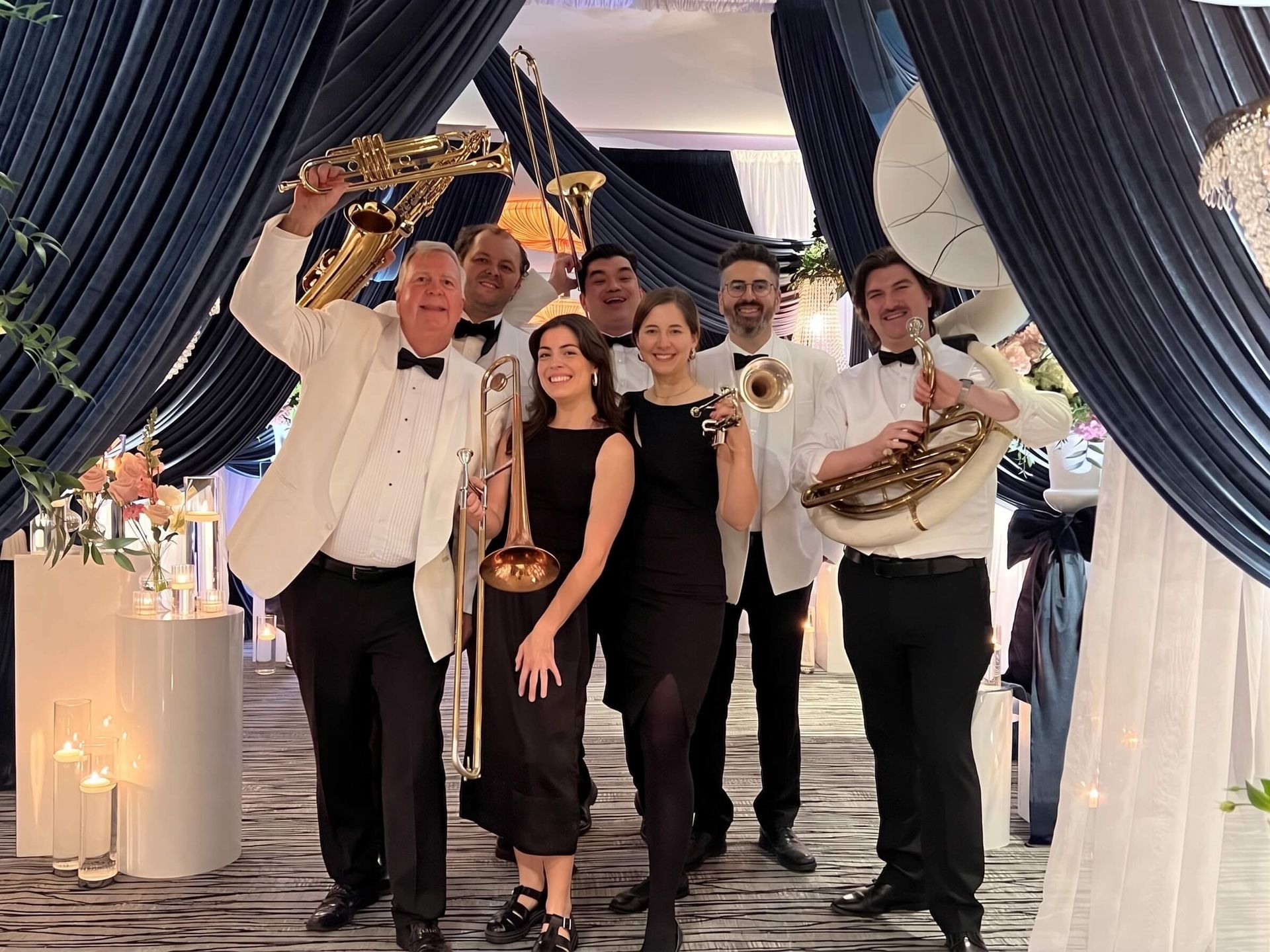 Brass band posing under dark drapes at a decorated event entrance, holding instruments and smiling