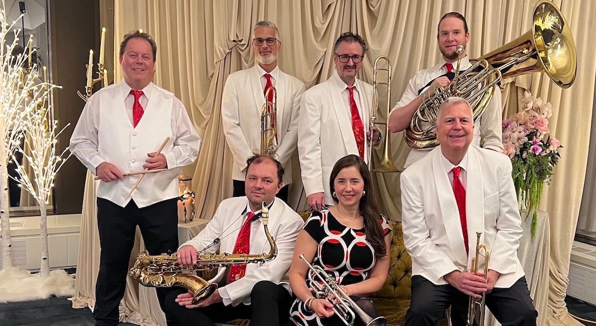 Group of musicians in white shirts and red ties posing indoors with brass instruments and flowers