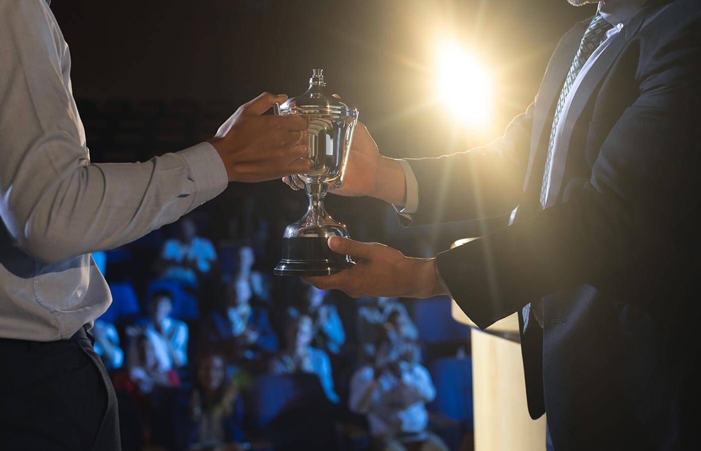 Two people clink glasses on a stage, with bright spotlight and audience in the background.