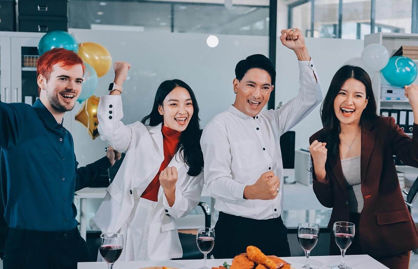 Four coworkers cheer at a table with balloons, smiling and raising drinks in a bright office setting