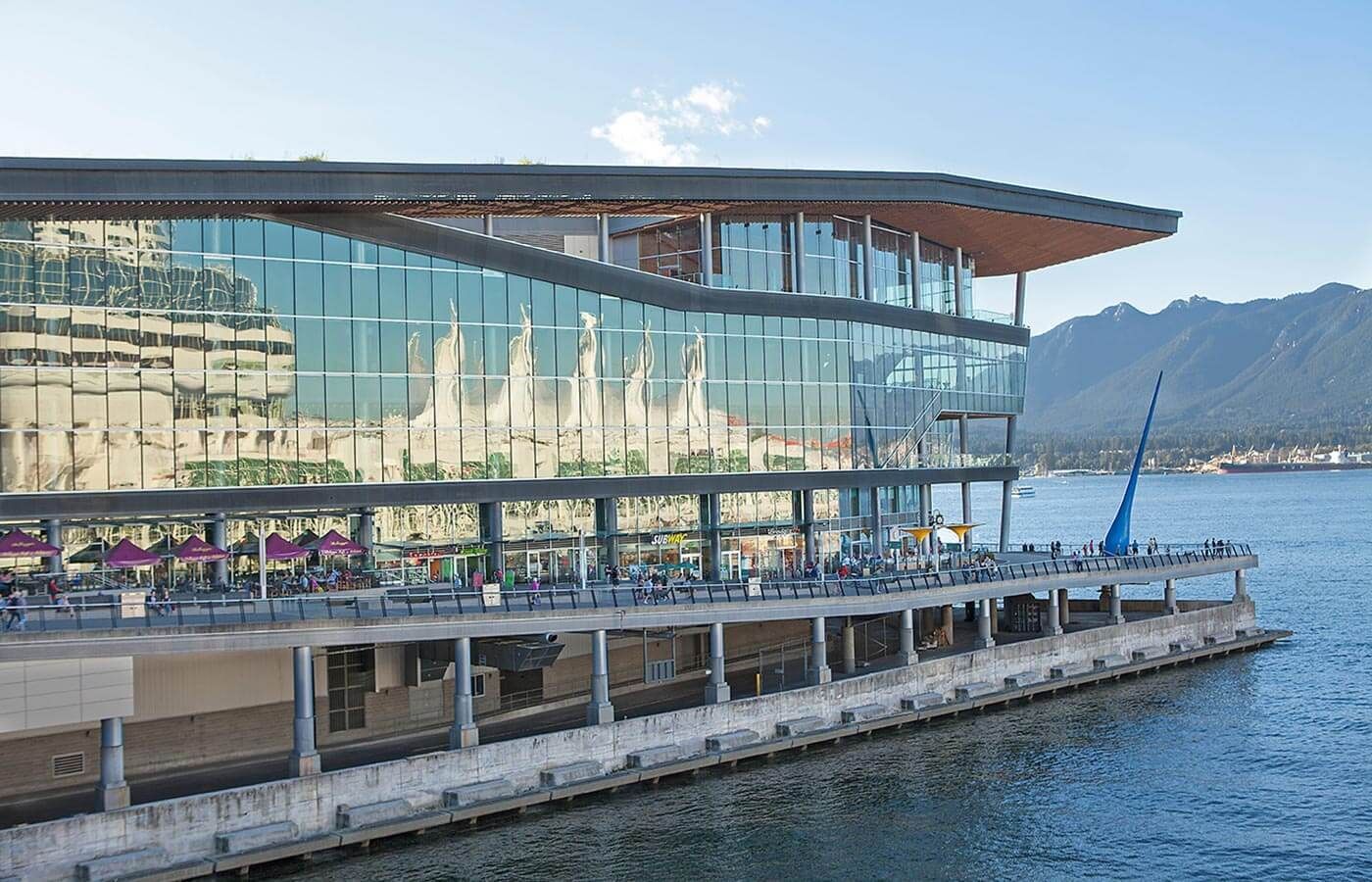 Modern glass waterfront building with a sloped roof beside a calm bay, backed by mountains