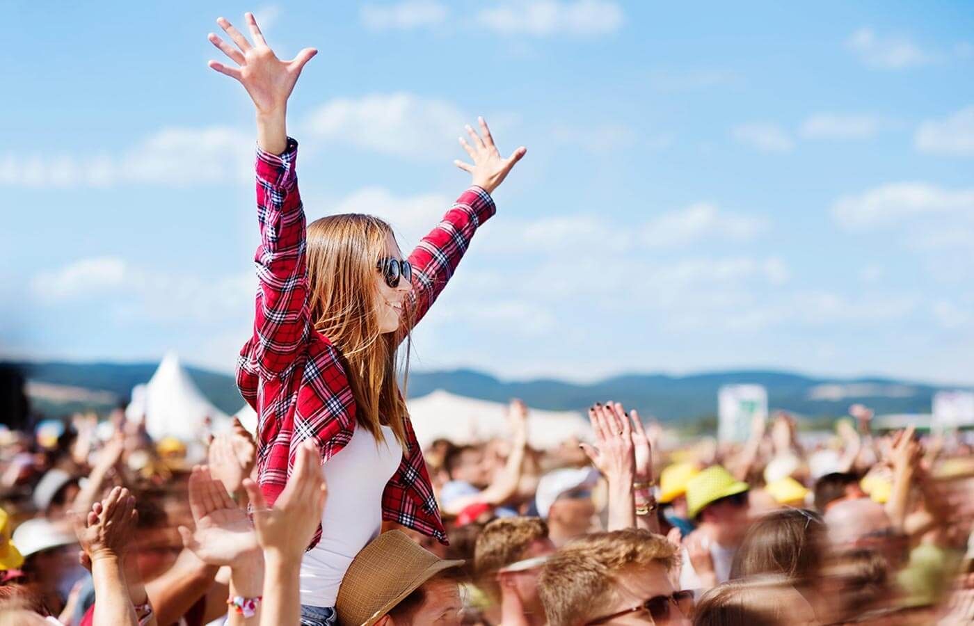 Crowd at an outdoor music festival, with a child on someone’s shoulders raising both arms under a blue sky