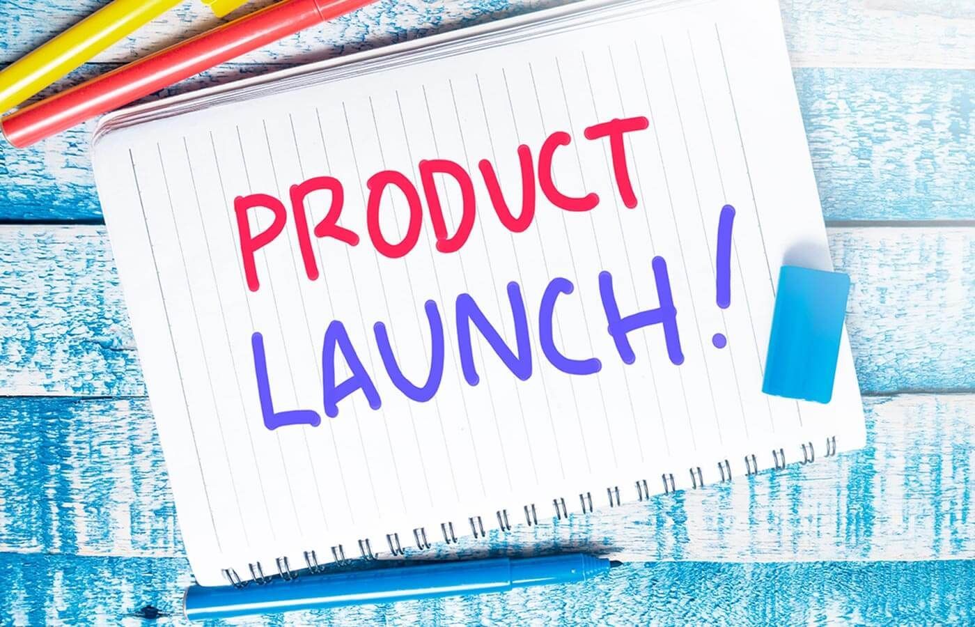 Notebook page with colorful pencils and handwritten “PRODUCT LAUNCH!” text on a blue wooden background