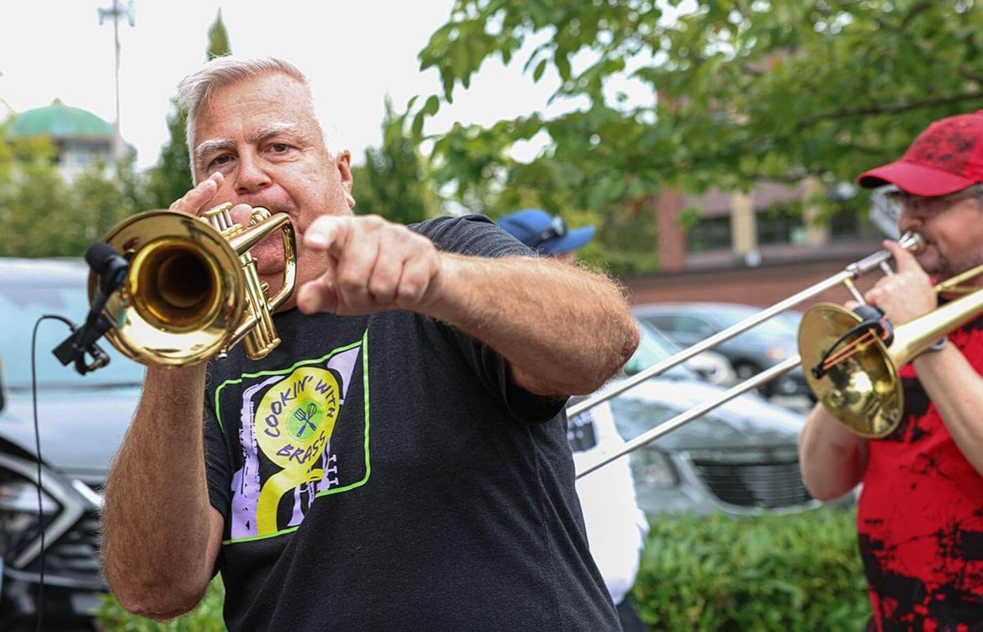 Two musicians playing trombone outdoors, one in a black shirt and one in a red cap.