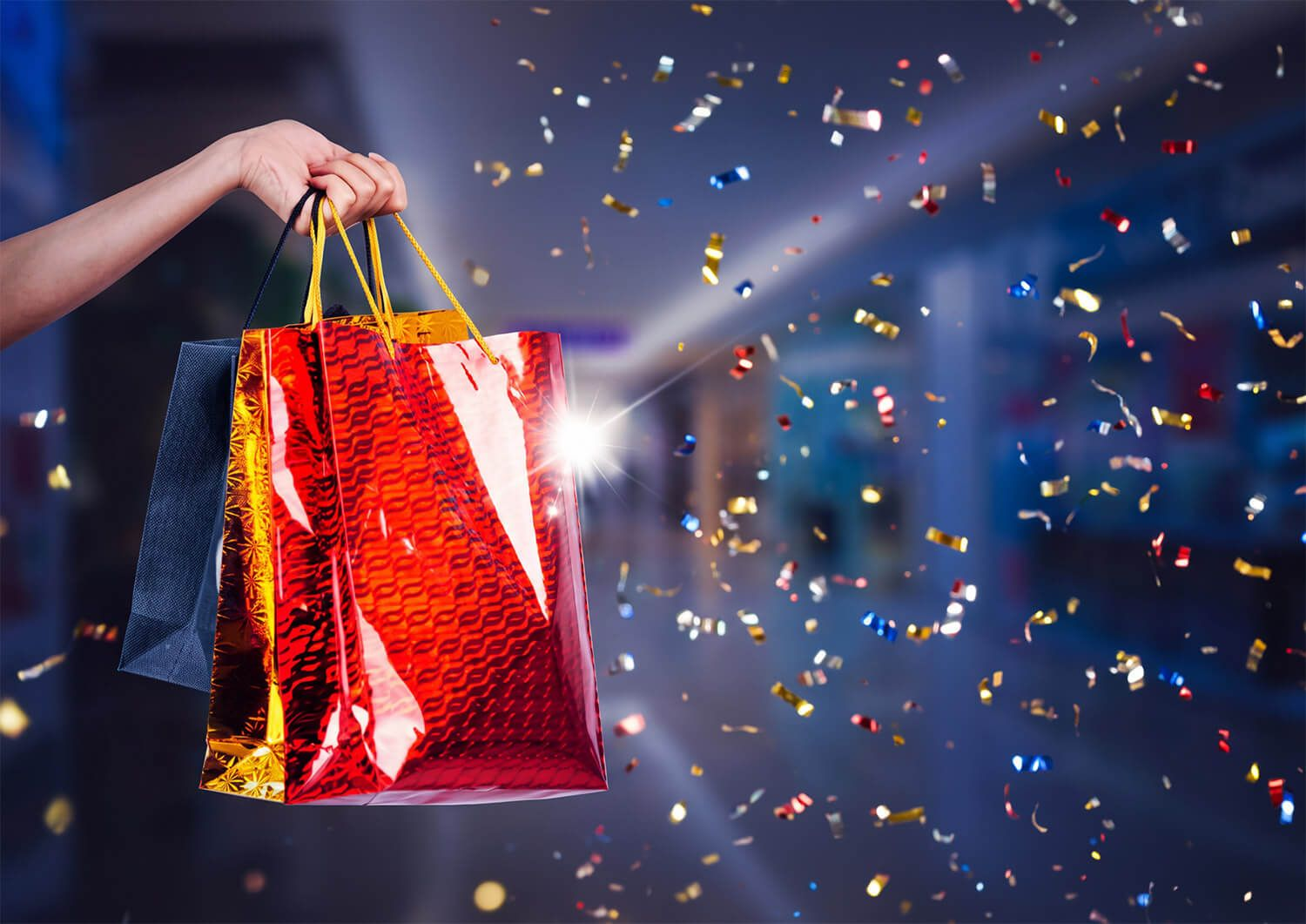 Hand holding a shiny red gift bag with confetti flying against a dark blue background