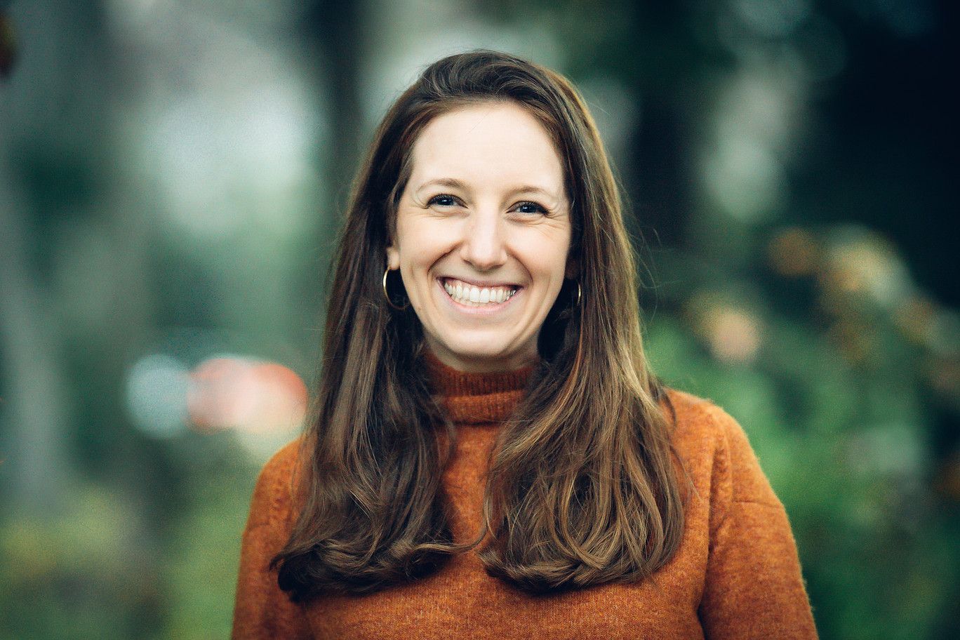 Smiling person in a rust-colored sweater against a blurred outdoor background
