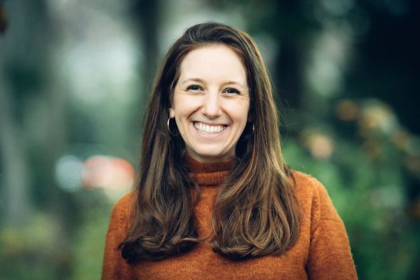 Smiling woman in an orange sweater standing outdoors with a blurred green background