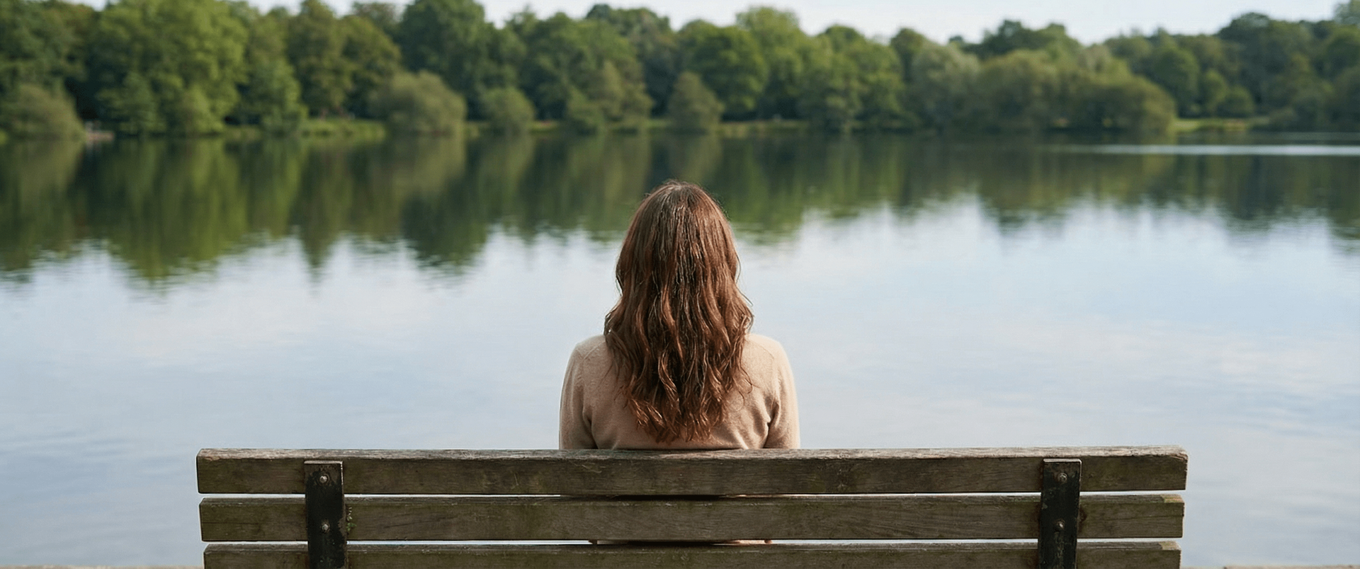 Person with curly hair sits on a bench facing a calm lake and tree-lined shore.