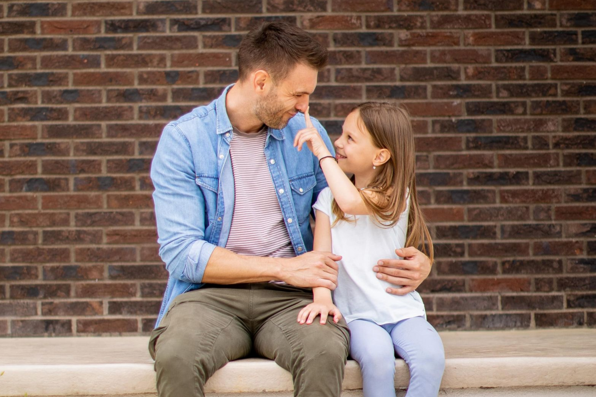 Adult and child sitting on steps, smiling and touching faces in front of a brick wall.
