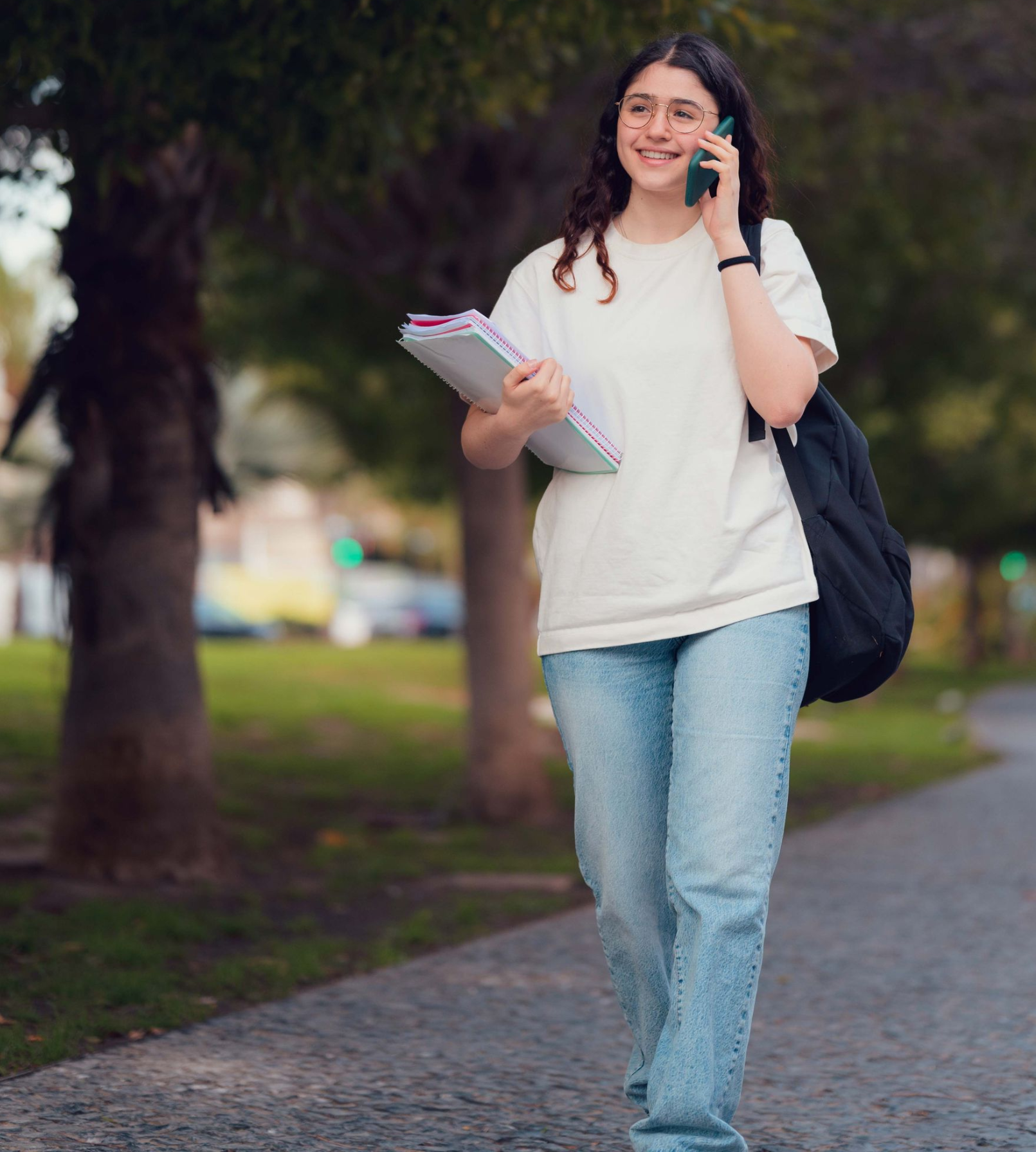 Young woman walking on a park path, smiling and talking on a phone, carrying notebooks and a black bag.