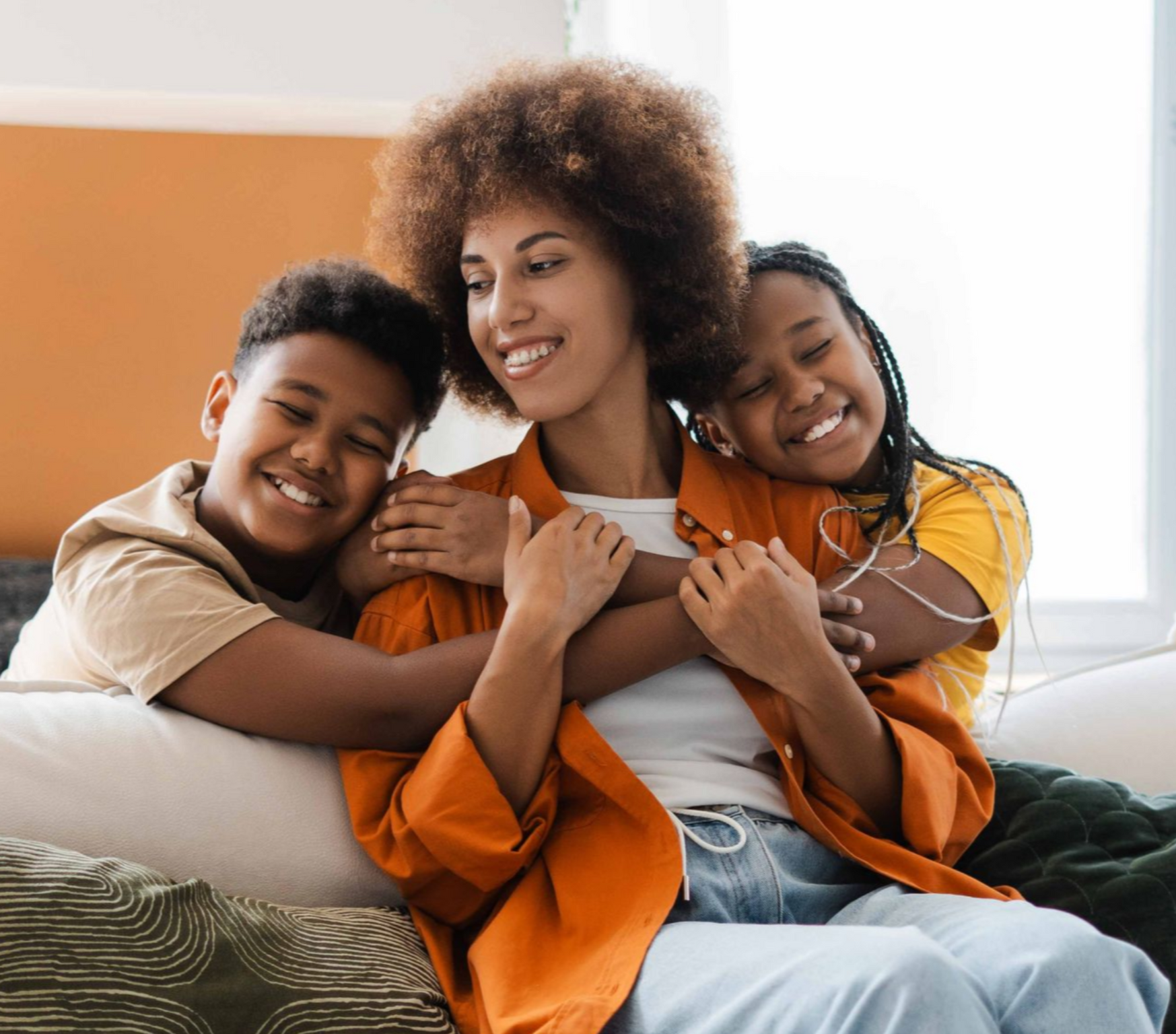 Three people smiling and hugging on a couch in a bright living room, wearing orange and white clothing