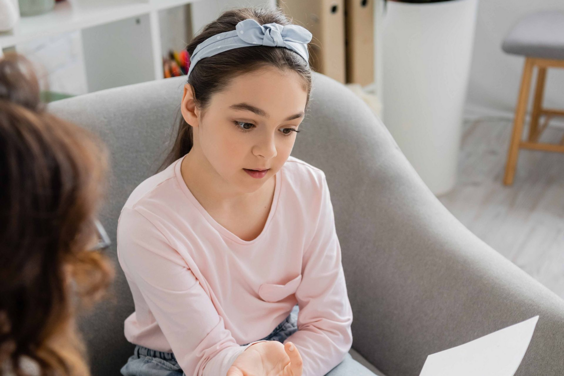Woman in a pink top sitting on a couch, talking with someone in a bright living room.
