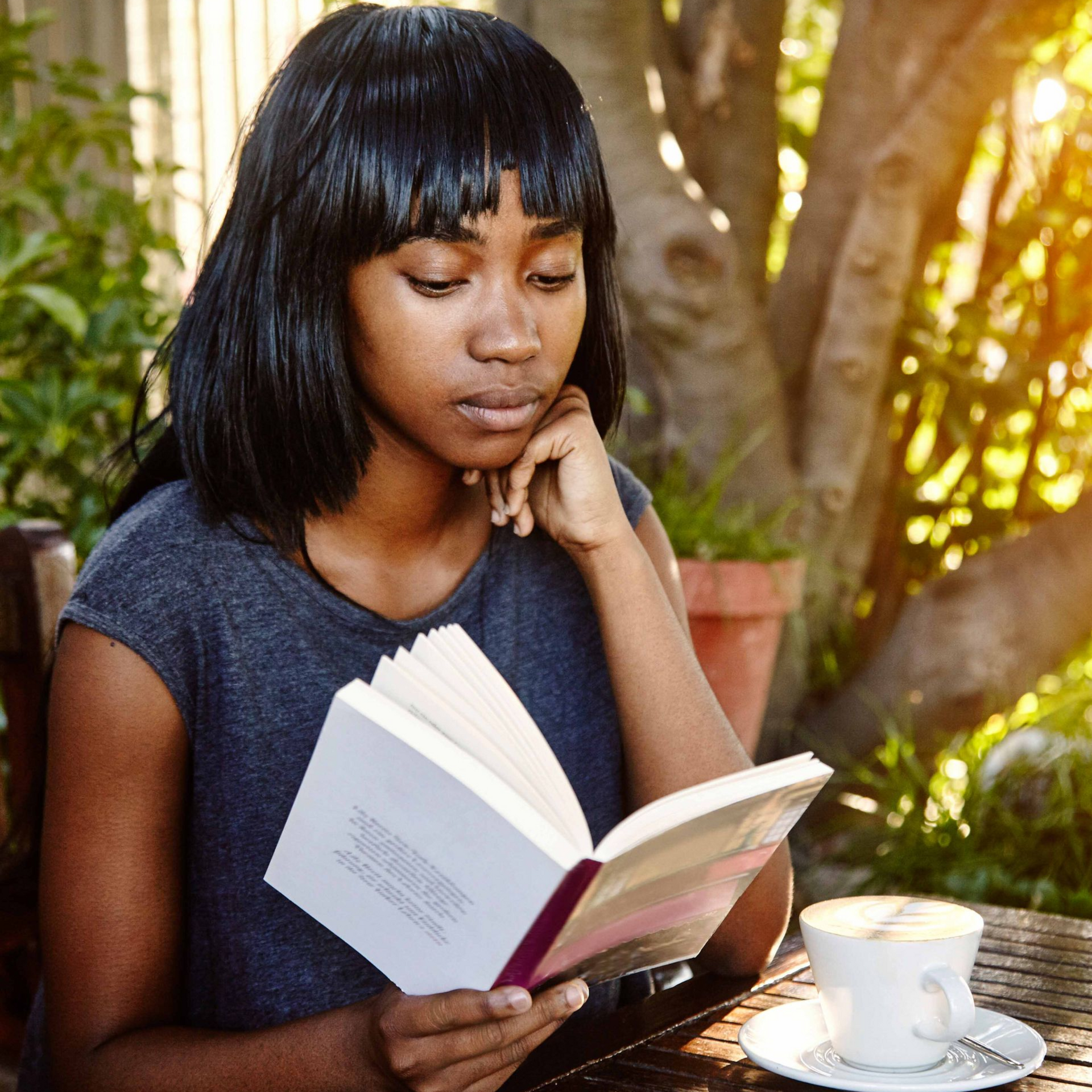 Woman reading a book at an outdoor table with a white teacup in a garden setting