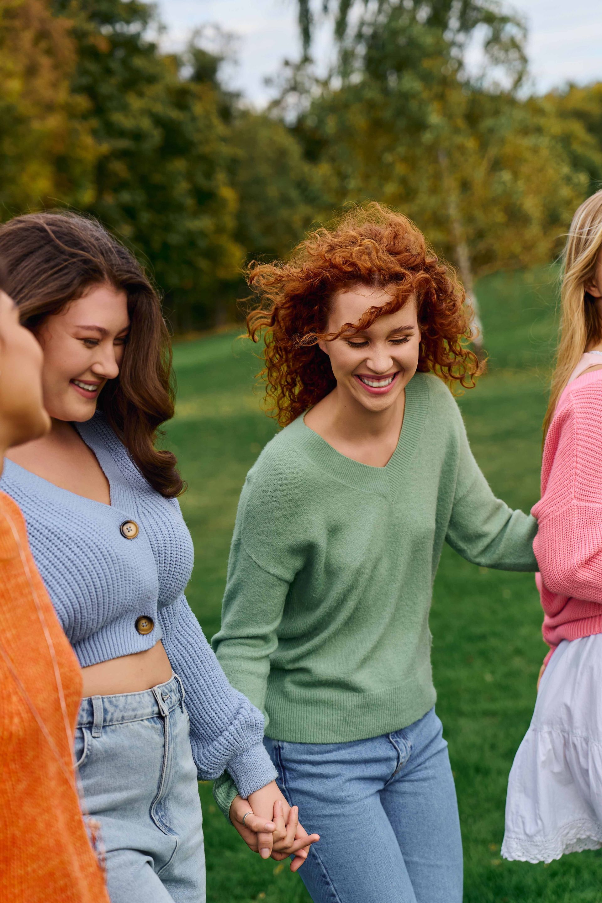 Three friends smiling and holding hands outdoors in colorful sweaters and jeans.