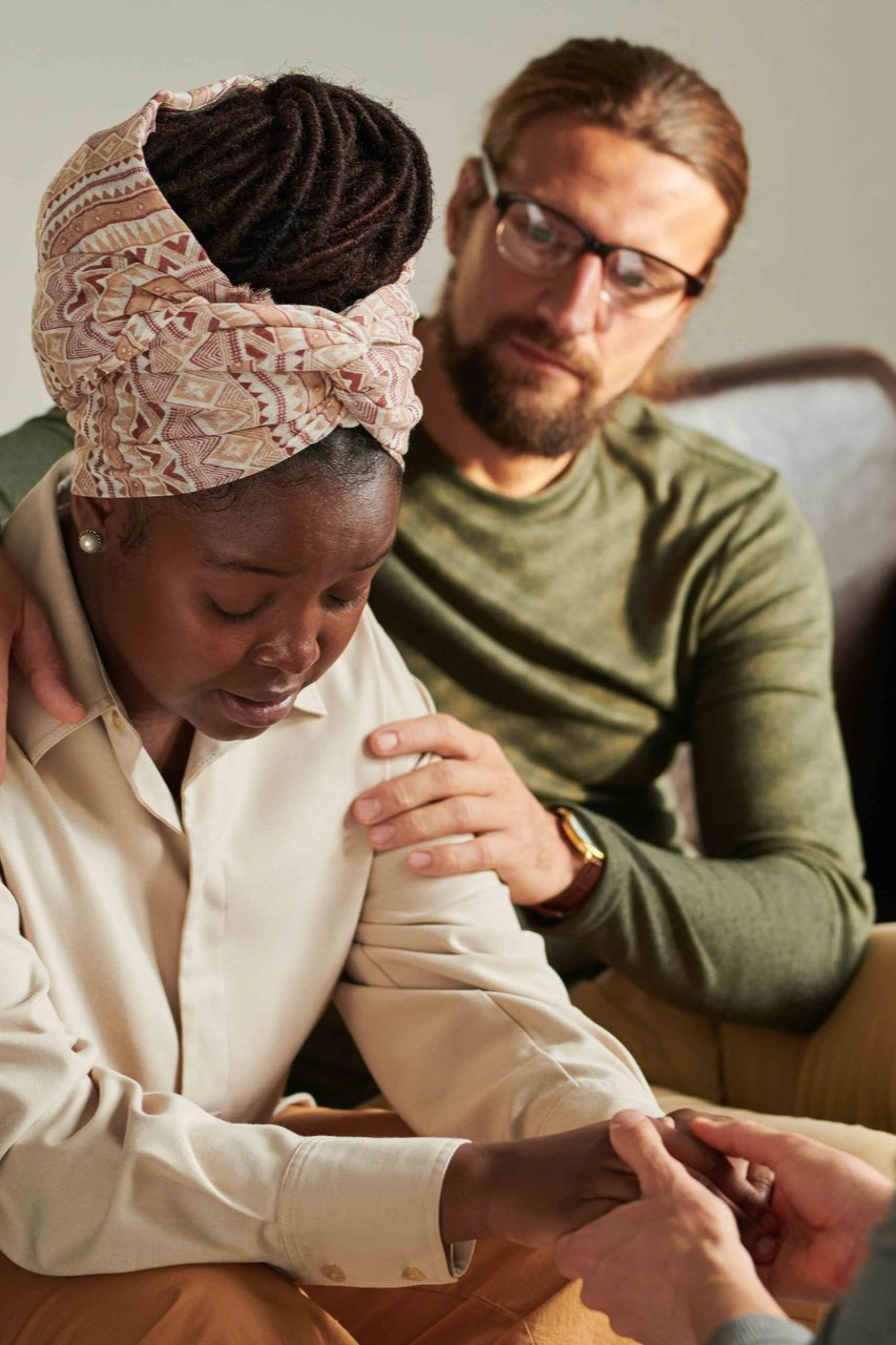 Two people sit closely, one comforting the other by holding her hand and shoulder indoors.