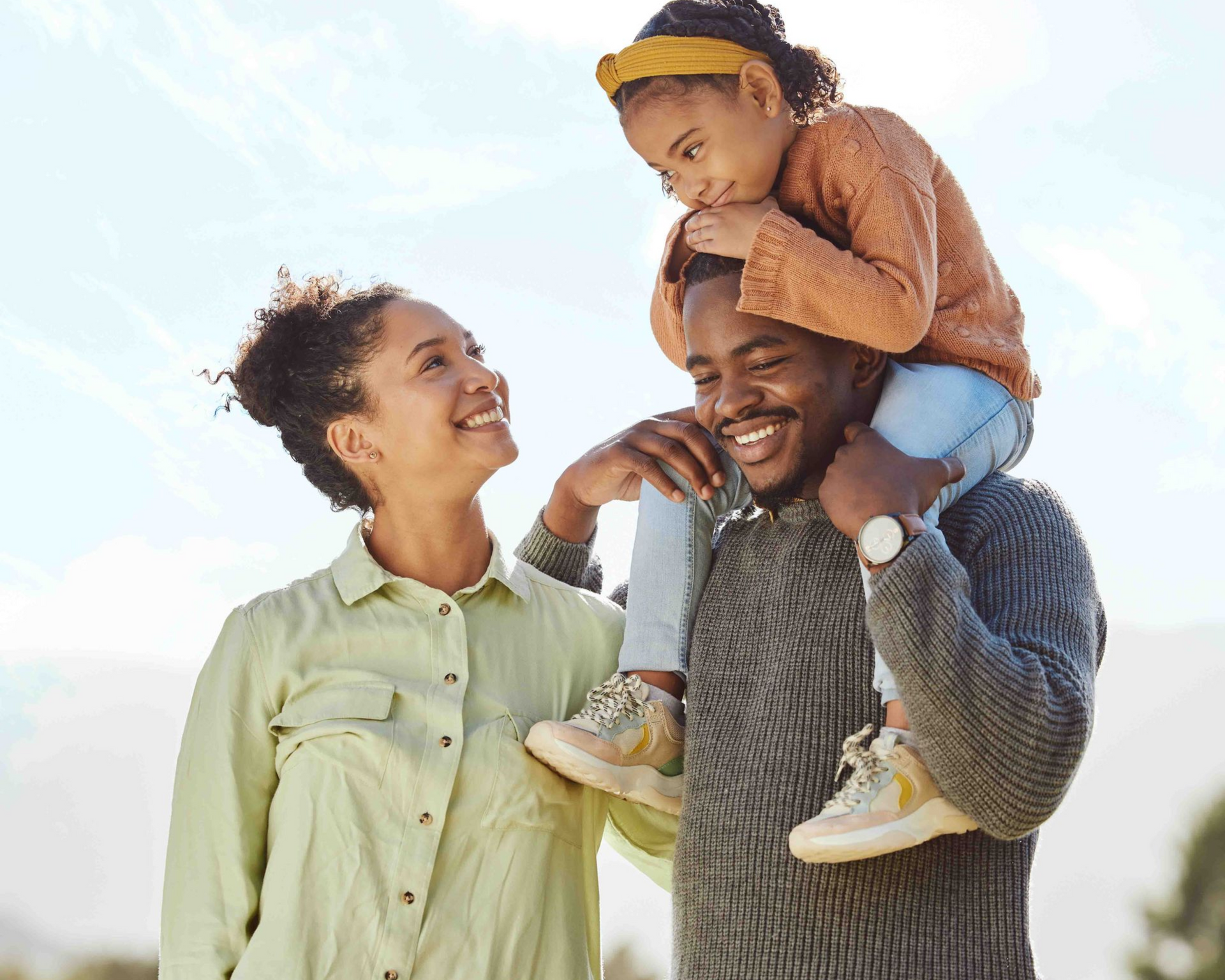 Two smiling adults outdoors, one carrying a child on his shoulders under a bright sky