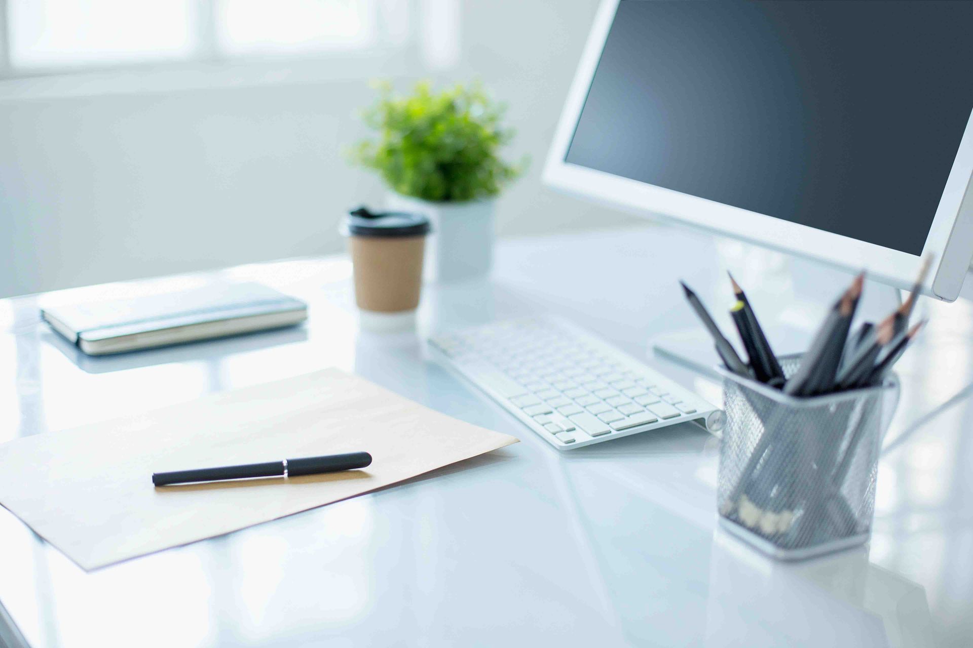 Minimal white office desk with monitor, notebook, pen, keyboard, and pencil cup on a bright background
