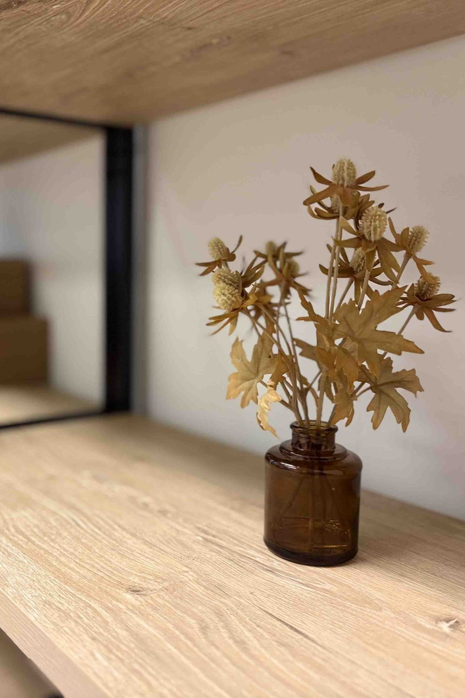 Small dried flower arrangement in a brown glass jar on a wooden shelf against a white wall.