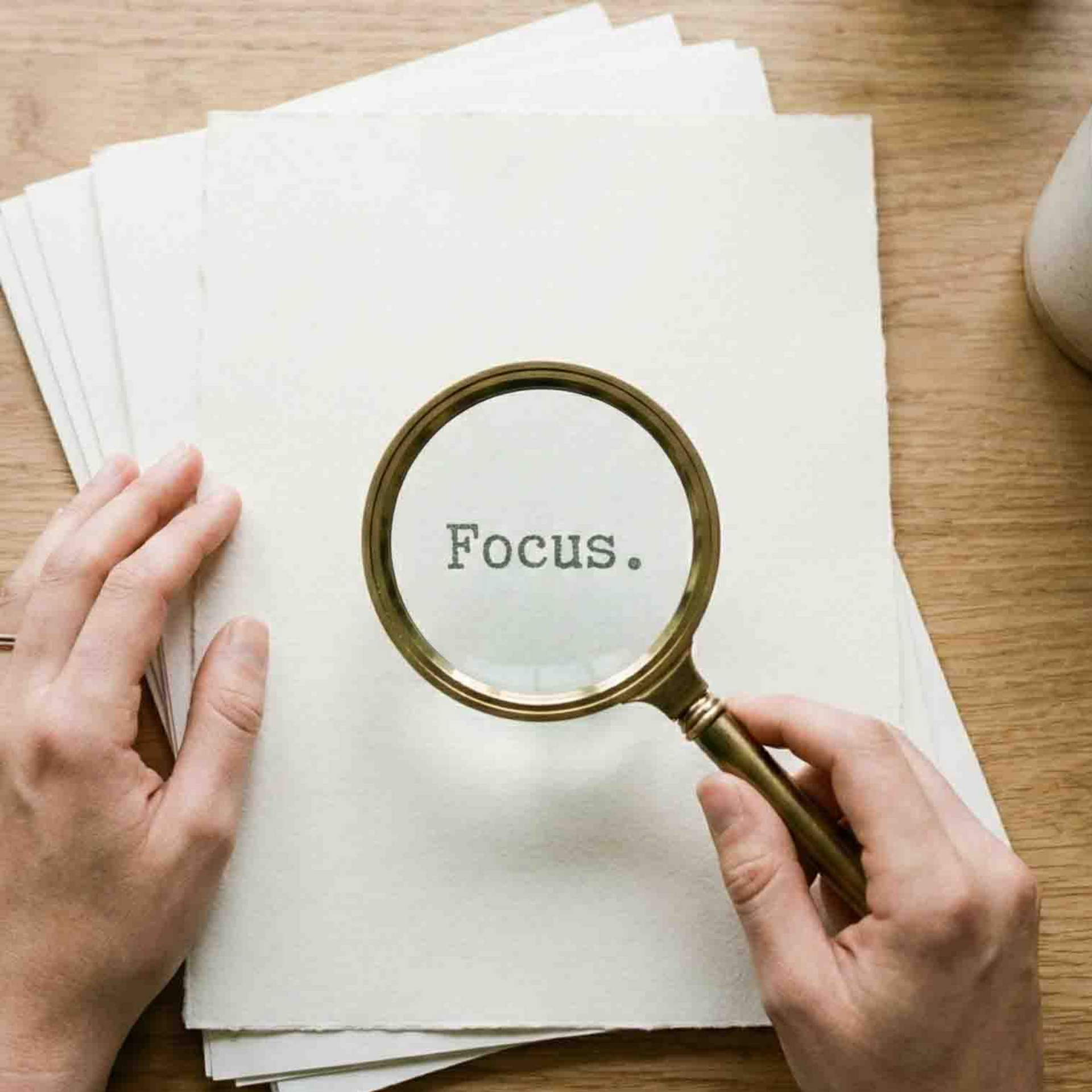 Hands holding a magnifying glass over paper with the word “Focus.” on a wooden desk