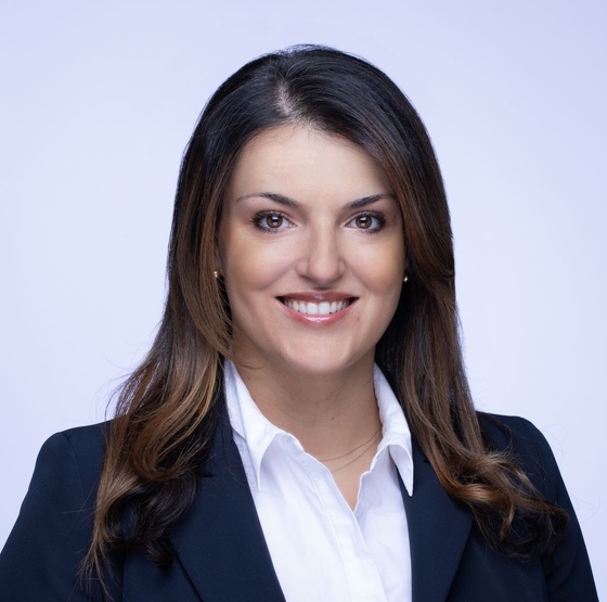 Woman in a blazer and white shirt smiles at the camera against a light blue background.