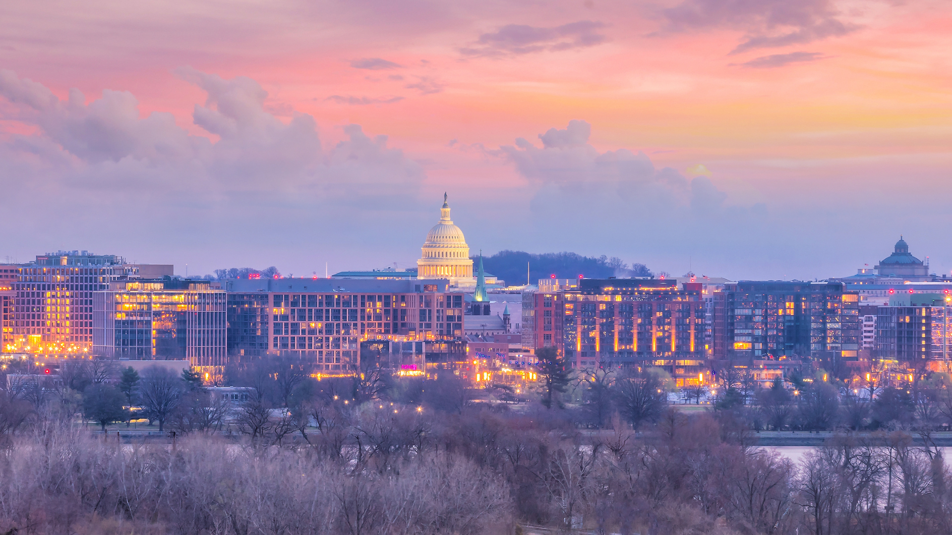 City skyline at dusk with a lit domed capitol building beneath pink and purple clouds