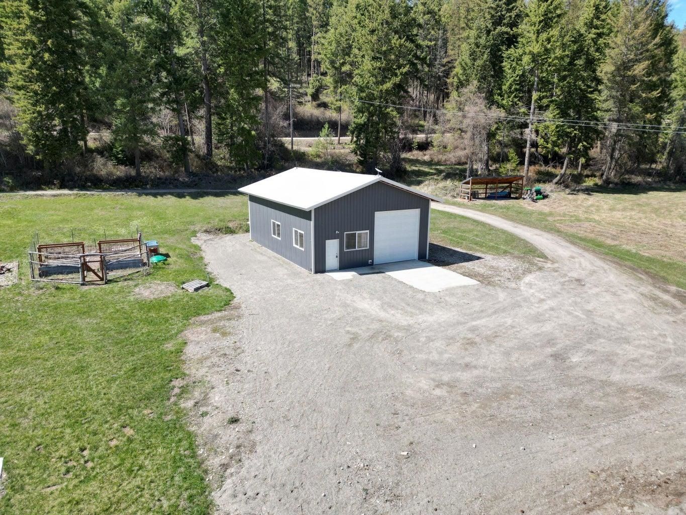 Small gray shed with white roof on a gravel clearing beside a grassy yard and forest trees.