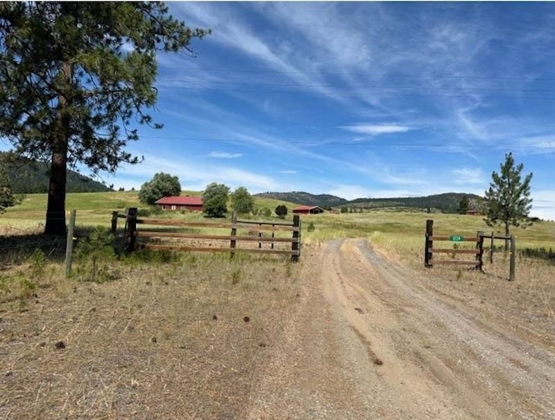 Rural dirt road through open ranch gate, with red barn, green fields, trees, and blue sky.
