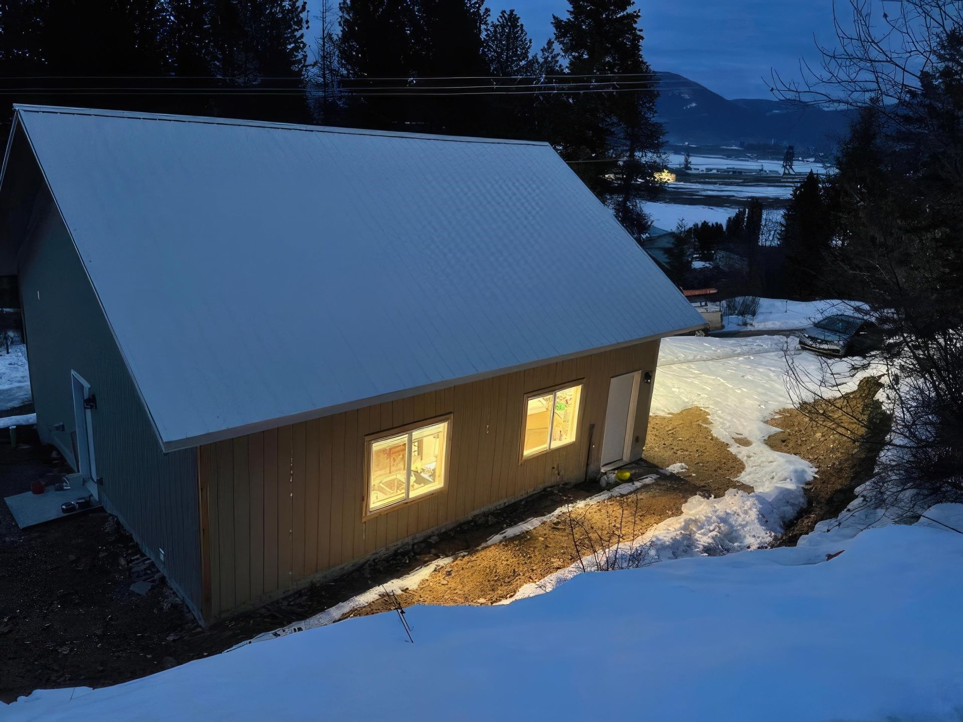 Snow-covered cabin at dusk with warm lit windows in a wooded landscape