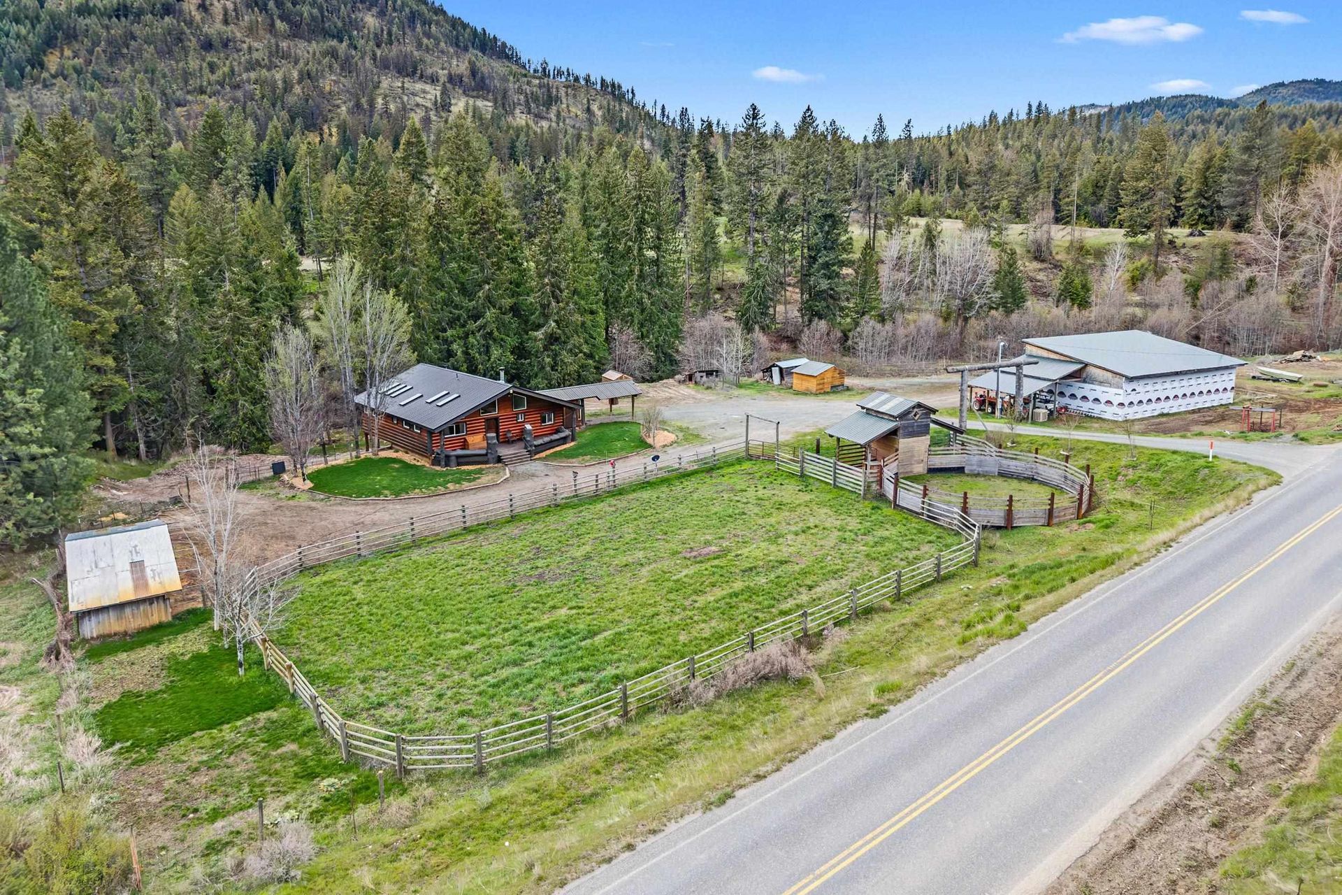 Rural homestead with green garden plots, barns, and a road beside forested hills