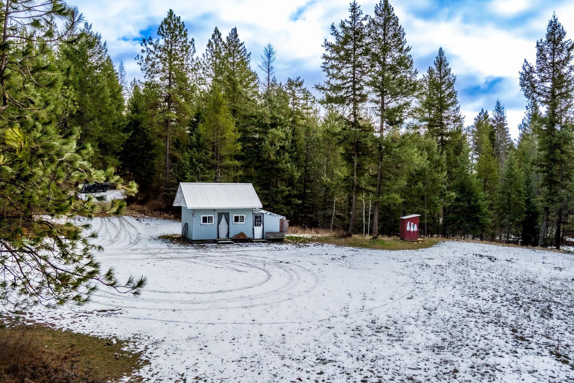 Snow-covered clearing with a small white cabin and a red shed among evergreen trees under a cloudy sky